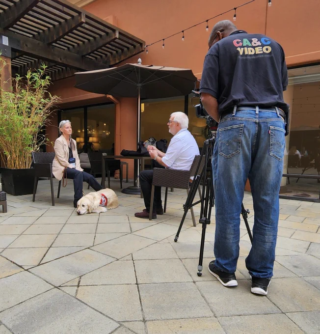 Mom is sitting in a chair wearing a tan and b lack business suit. Sampson is lying at her feet wearing a red service dog vest. They are being interviewed by a man, and a videographer is recording the interview. 