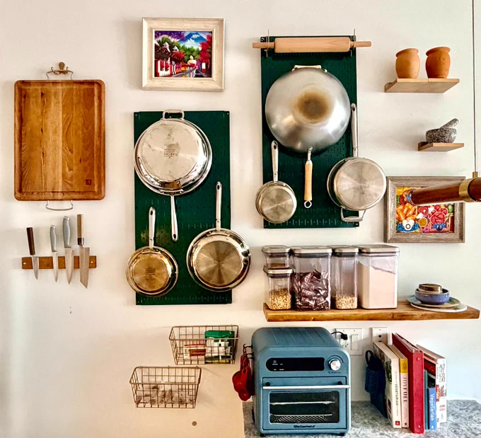 Organized kitchen wall featuring dark green Wall Control metal pegboards holding stainless steel pots, pans, and utensils. The setup includes floating shelves, wooden cutting boards, spice containers, cookbooks, and a compact countertop oven, creating a clean and functional modern kitchen workspace.