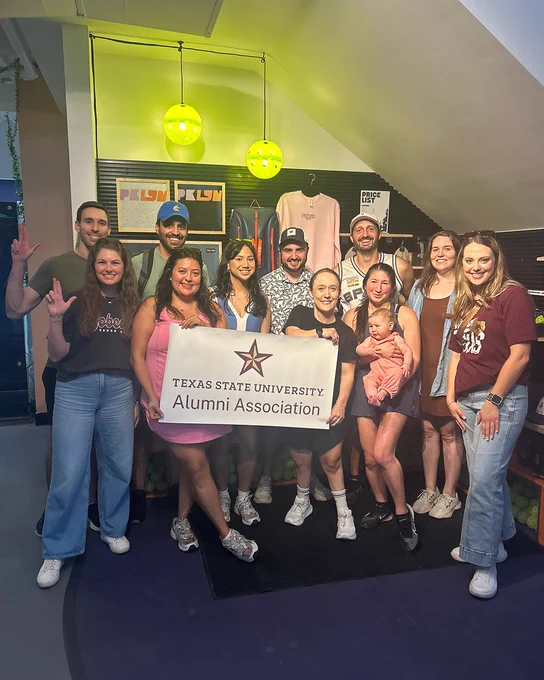 Group of alumni at a pickleball court holding up an Alumni Association banner  