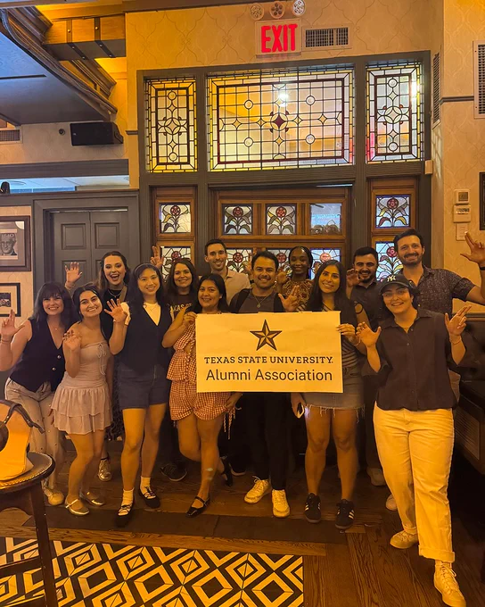 Group of alumni in a dimly lit restaurant bar in New York holding up an Alumni Association banner  