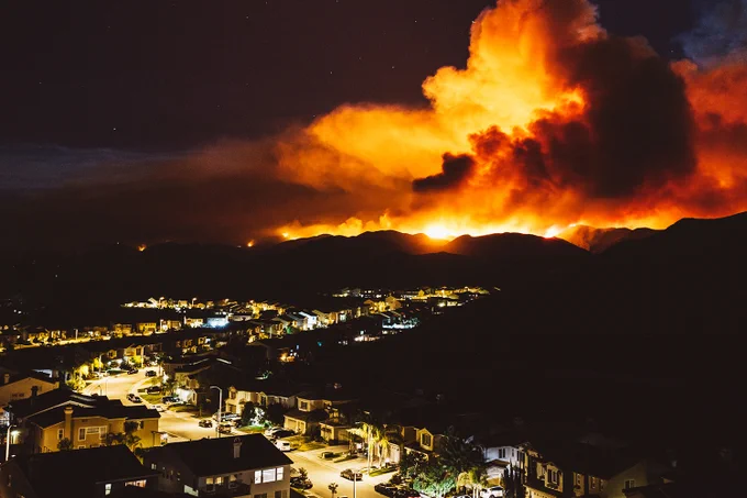 IMAGE : Photo d'un quartier la nuit, sous un feu de forêt qui embrase la colline, avec des flammes orange vif et une épaisse fumée qui illuminent le ciel.