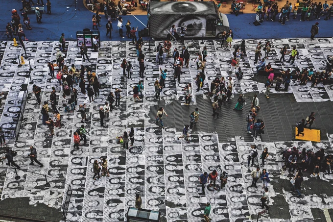 Crowd gathered in a large public square covered with a striking black-and-white artwork made up of hundreds of portrait faces arranged in a grid. People walk, stand, and gather across the installation, while a nearby structure displays a large close-up face image. The scene is viewed from above, showing the scale of the artwork and the busy atmosphere.