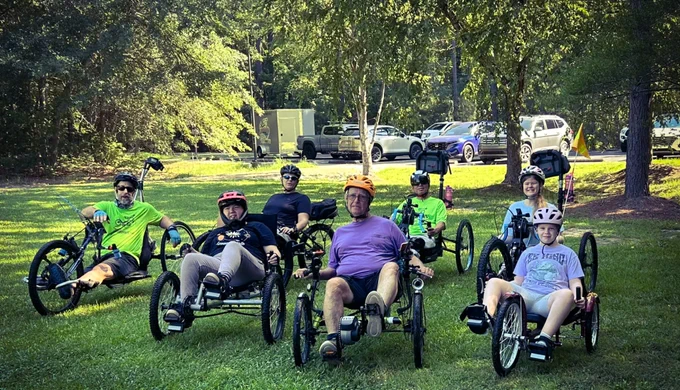 Group of seven people sitting on recumbent trikes on a grassy area surrounded by trees and parked cars in the background.