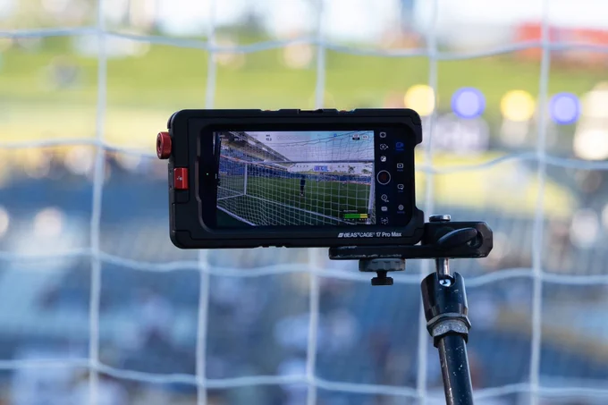 An iPhone 17 Pro is horizontal in the center of the frame. A soccer field is visible on the screen of the iPhone, displaying the view from the camera. Behind the iPhone, a soccer net and stadium are visible but out of focus.