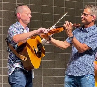 Two men performing on stage. They are holding and playing guitars and violins, helping each other to play their instruments.

