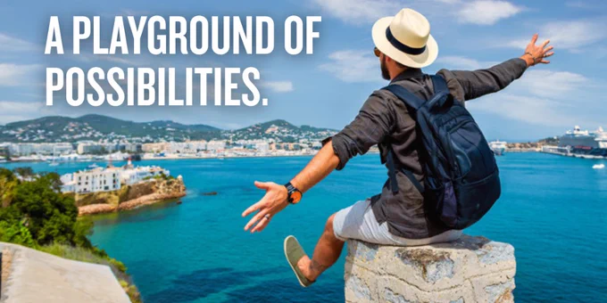 Man wearing a hat and backpack sits on a rock overlooking a vibrant coastal city and harbor under a blue sky.