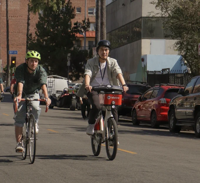 Two cyclists ride side by side on a city street lined with parked cars and buildings under clear daylight.