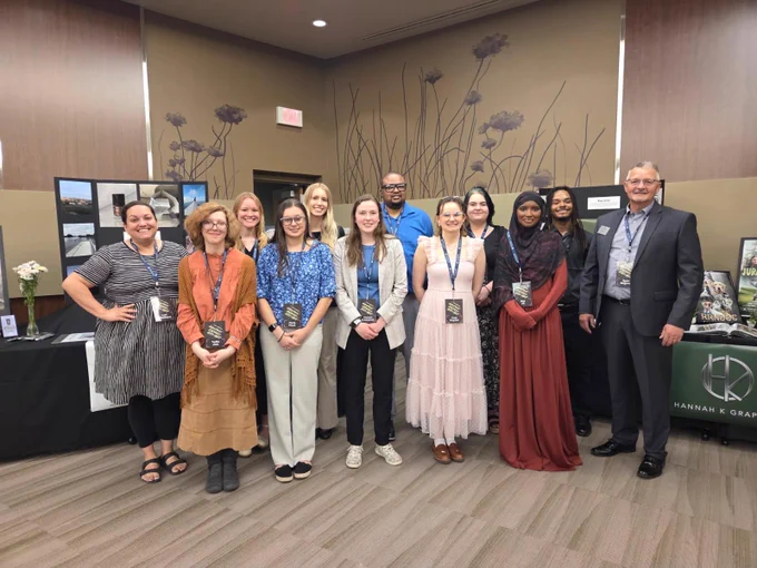 A group of eleven people are standing together at an indoor event or conference. They are all smiling and wearing name tags. There are tables in the background displaying materials and promotional items from different organizations. The room is decorated with large illustrations of flowers on the walls.