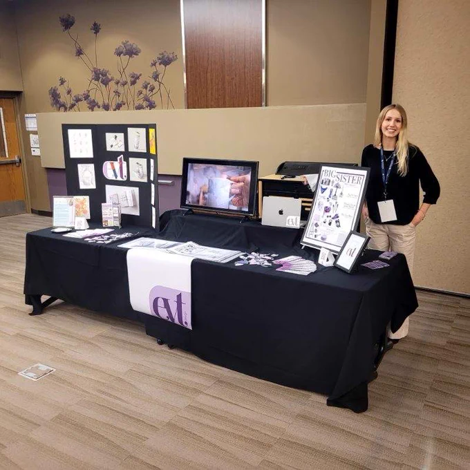 A person stands beside a display table at an event. The table features various printed materials and a screen showing a video. There is a sign on the table that reads "Big Sister." The setup includes design elements, such as illustrations and logos, on black and purple tablecloths. The background has floral wall art.