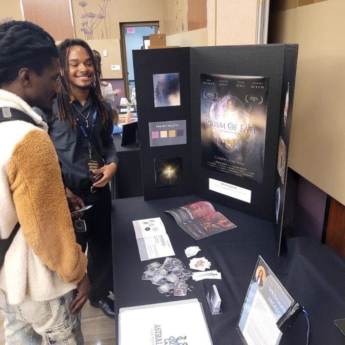 Two individuals are standing by a tabletop display for a project titled "Prism of Fate" at an event. The display includes images, text, and brochures. They appear to be engaged in conversation, and promotional materials are spread across the table. The room has a beige and cream color scheme.