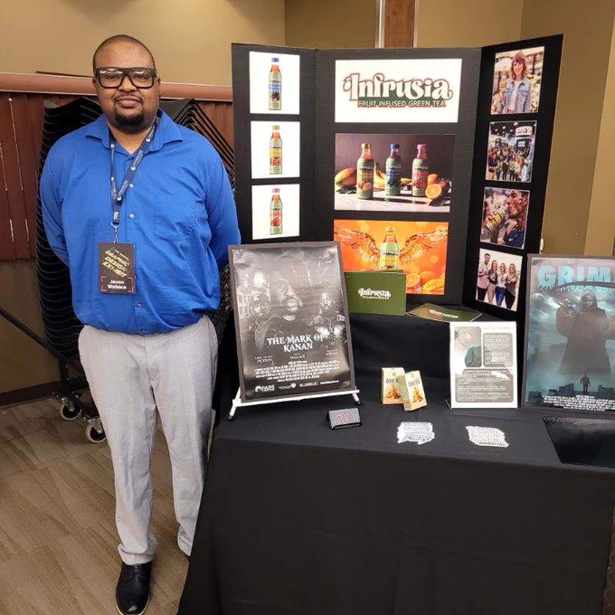 A person stands next to a display table featuring products and promotional posters, including "Infusia" beverages and the movie "The Mark of Kanan." The backdrop includes various promotional images. The person is wearing a blue shirt and gray pants with a badge around their neck.