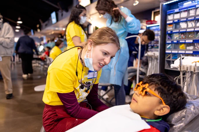 A dental student sits and talks to a young child laying in a dental chair. The child wears star-shaped sunglasses.