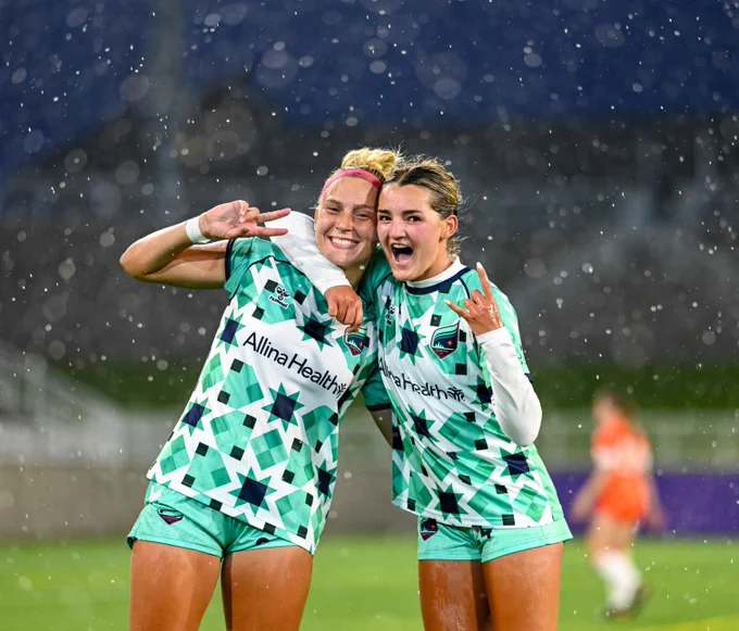Two soccer players in matching green and white star-patterned jerseys with "Allina Health" logo are posing happily on a rainy field. They are both making hand gestures and smiling, with blurred spectators and players in the background.