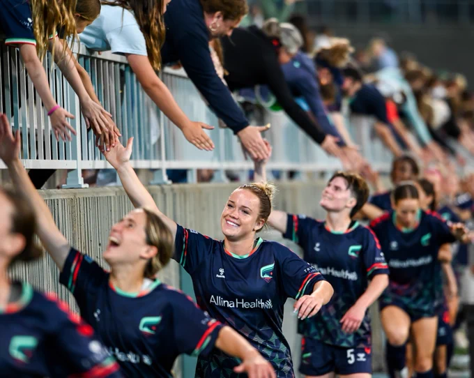 Soccer players in navy blue jerseys with the Allina Health logo high-five fans over a railing. Excited supporters reach down as the players celebrate.