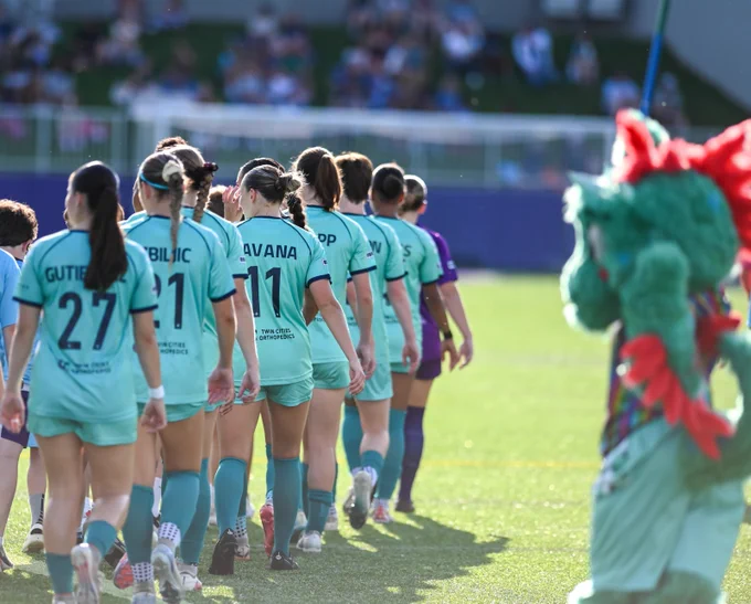 A sports team in teal uniforms is walking in a line on a soccer field. Names and numbers are visible on the back of their shirts. A person in a green dragon costume is on the right side. Spectators can be seen in the blurred background.