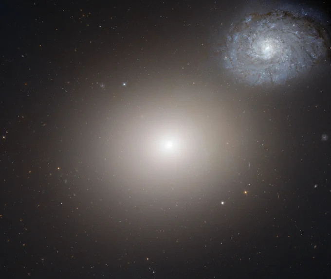 A hazy elliptical galaxy shines at image center, with a blue-white spiral galaxy appearing smaller at upper right.