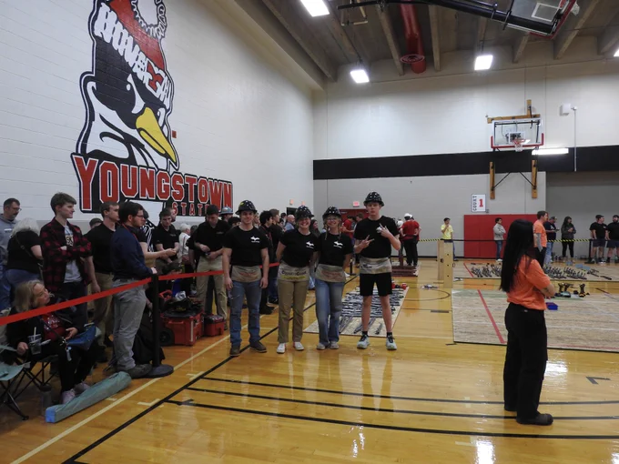Members of Michigan Tech’s Steel Bridge Team stand on the competition floor inside a gymnasium at Youngstown State University during the ASCE Eastern Great Lakes Regional Competition. Team members wearing black shirts and hard hats pose in front of bridge components and tools while spectators and other competitors gather nearby. A large Youngstown State Penguins logo is painted on the wall in the background.