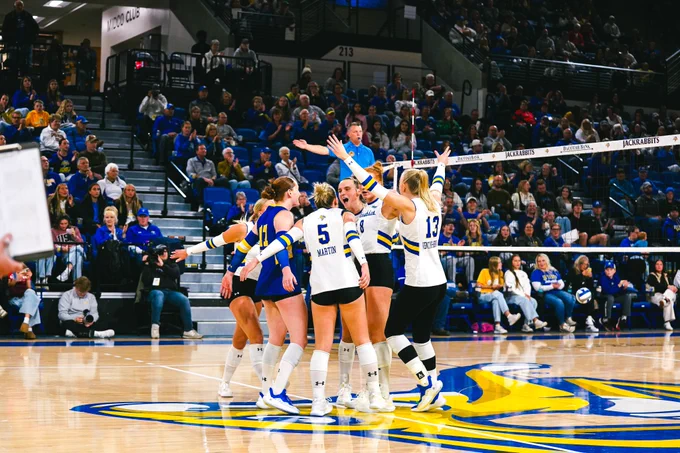 SDSU volleyball players wearing white and blue uniforms gather at center court on a Jackrabbits logo, raising their arms and forming a tight huddle near the net, with “Jackrabbits” visible on the net tape and a full crowd seated in the arena stands behind them.