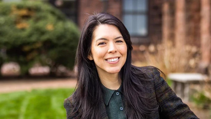 The image shows a person with long dark hair, smiling outdoors. The background includes blurred greenery and a building.