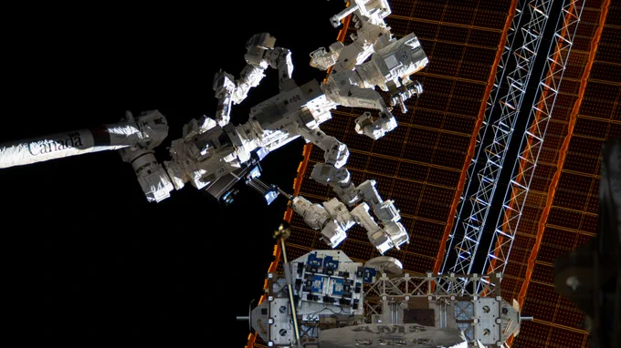 A large robotic system with dual arms and several joints is shown extended above external hardware outside of the space station. The robotic arms are white against the blackness of space, and the station’s large orange solar arrays fill the right side of the frame. In the middle of the bottom of the image is sample hardware, shown as blue and black rectangles on a large white surface.  