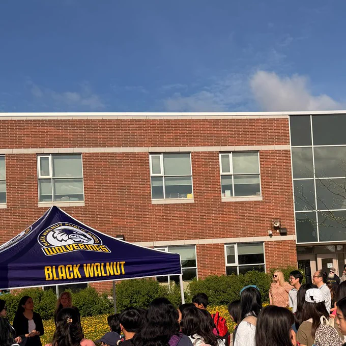 Crowd of students and adults gathered outdoors in front of a brick school building under a blue sky. A canopy tent labeled “Black Walnut Wolverines” is set up, and many students wear backpacks while facing speakers under the tent.