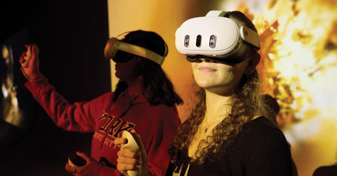 Two individuals wearing virtual reality headsets, one in a red sweatshirt with a "York University" logo, are interacting with VR controllers in a dimly lit environment.