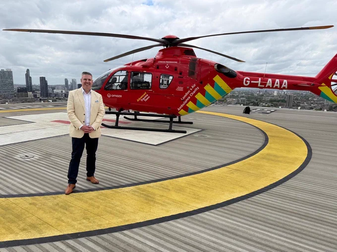David Holdsworth standing on a rooftop helipad in front of a red air ambulance helicopter.