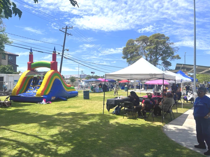 A lively outdoor community event features a colorful inflatable slide and several tents and tables set up on a grassy area. People are gathered under the tents and in the open space, enjoying the sunny day. The sky is clear with a few clouds.