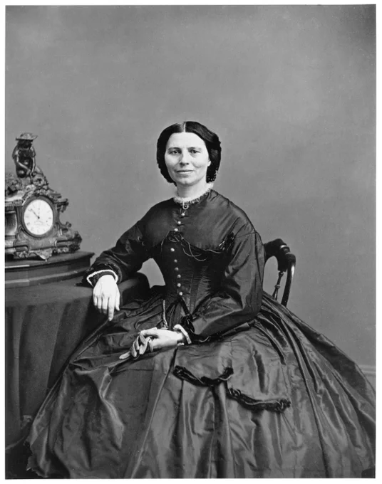 A historical portrait of Clara Barton seated beside an ornate clock. The individual is wearing a long, dark, Victorian-era dress with buttons down the front. The hairstyle is parted in the center and pulled back. The overall setting is formal and from a bygone era.