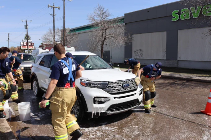 Edmonton Fire Rescue recruits wearing turnouts and blue t-shirts wash a white Ford SUV in a Save-on-Foods parking lot.