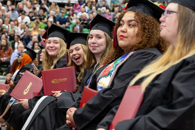 Gateway graduates in black caps and gowns seated in a row holding red diploma covers during a commencement ceremony.