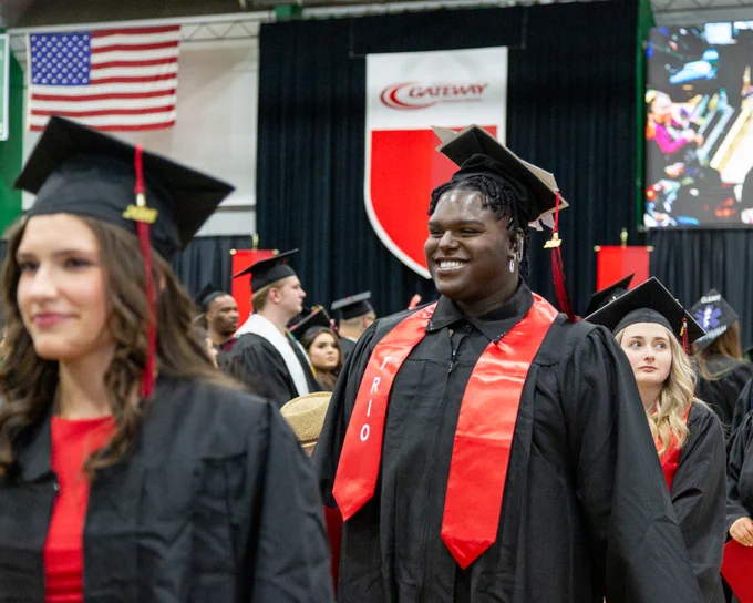 Gateway graduates in black caps and gowns with red stoles stand during a commencement ceremony, with an American flag in the background.