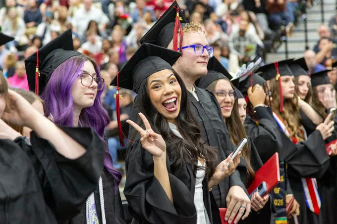 Group of Gateway graduates in black caps and gowns celebrating together at a crowded indoor ceremony.