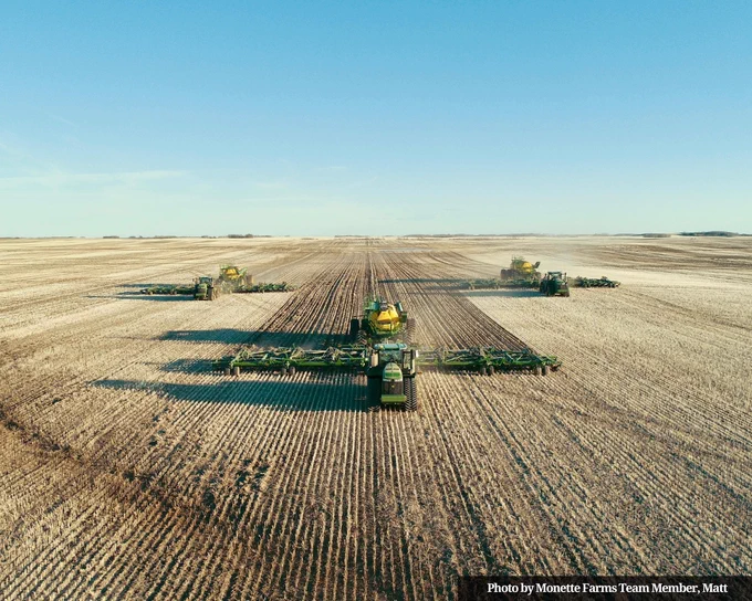 A wide drone shot of three sets of seeding machinary working in a row across a vast open field under a clear blue sky.