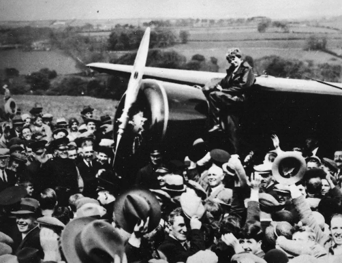 Photo of Amelia Earhart sitting on the nose of her Lockheed Vega airplane, surrounded by an enthusiastic crowd.
