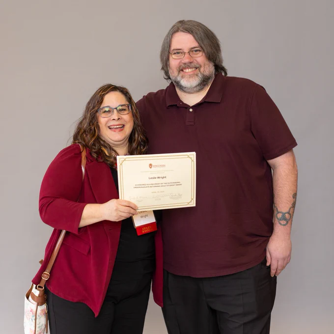Returning adult student Leslie Wright stands next to her husband holding a scholarship certificate, both are smiling warmly.