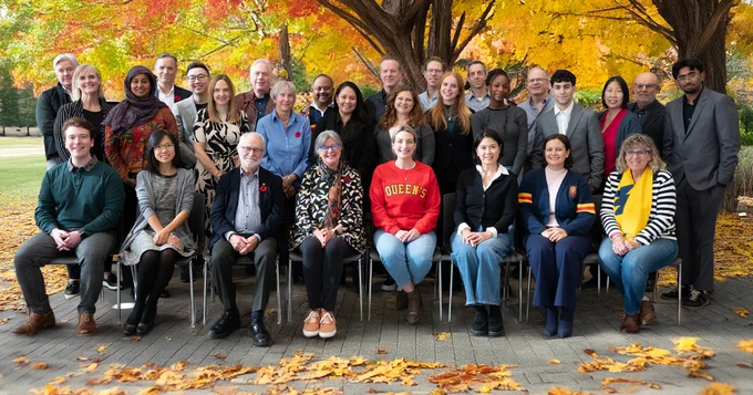 A group of people are gathered outdoors under trees. They are posing for a group photo, with some seated and others standing behind them. The setting is an outdoor area covered with fallen leaves. One person is wearing a red sweatshirt with "Queen's" printed on it.