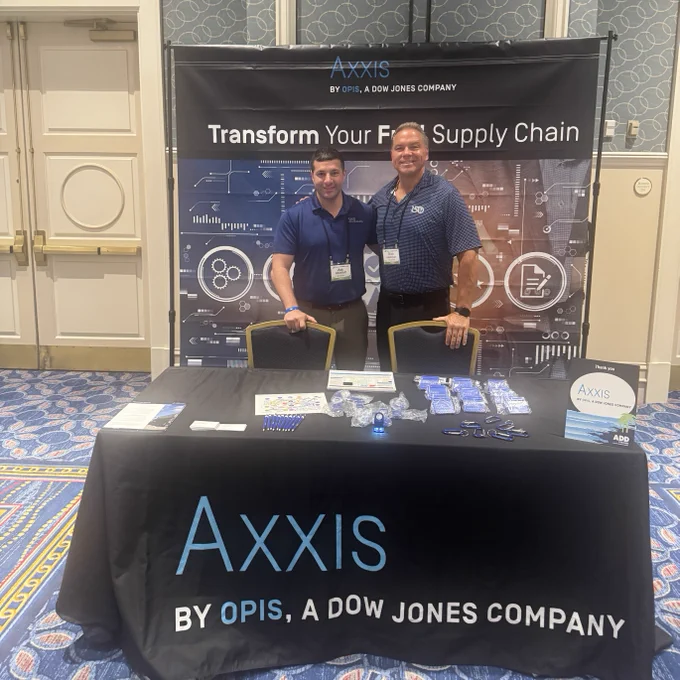 Two individuals stand smiling behind a table at an event. The table has promotional materials and displays a banner with "Axxis by OPIS, a Dow Jones Company," emphasizing supply chain solutions. The setting includes a patterned carpet and ornate wall decor.