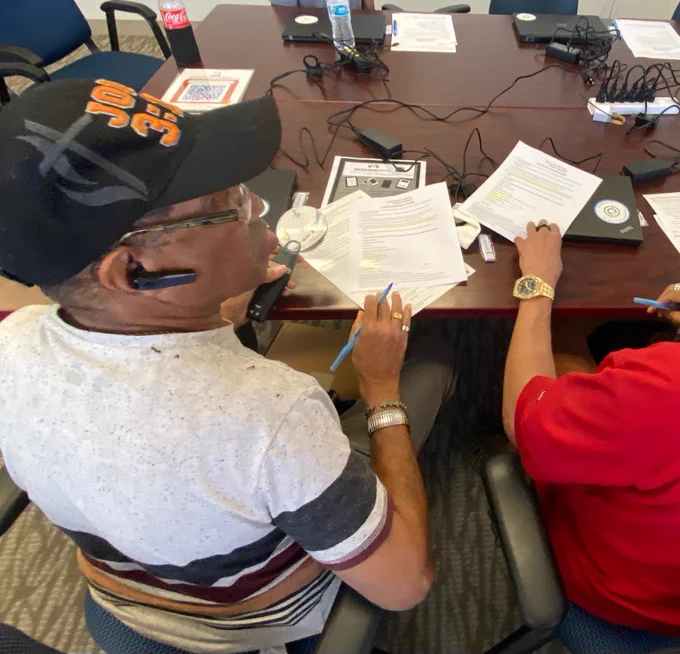 A person wearing a cap sits at a table with documents and a Coca-Cola bottle. There are multiple electronic devices and chargers on the table. Another person beside them is also involved with the documents.