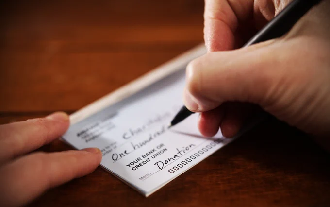 Hand writing a $100 donation amount on a personal check placed on a wooden table.
