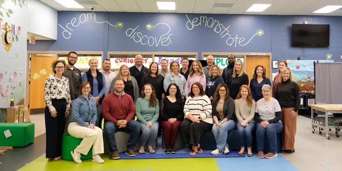 Group photo of 23 educators and staff posing in a colorful classroom with words 'dream,' 'discover,' and 'demonstrate' on the wall behind them.