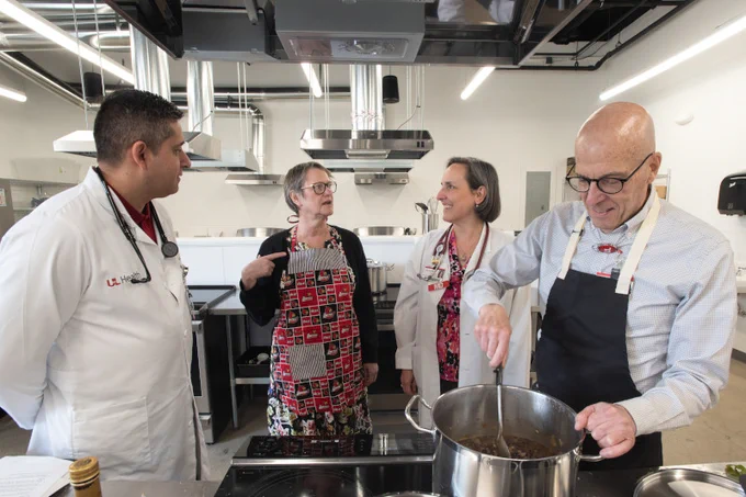 Four medical professionals in a kitchen setting, one stirring a pot while others observe and discuss cooking techniques.