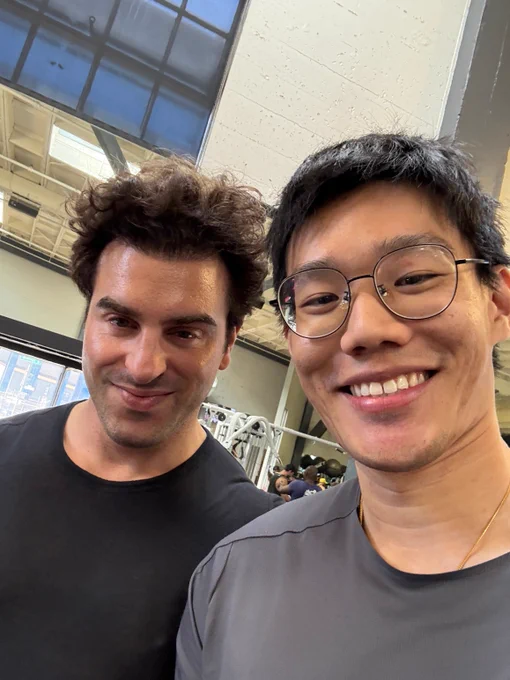Two men smile for a selfie in a gym, showcasing the workout space and equipment in the background.