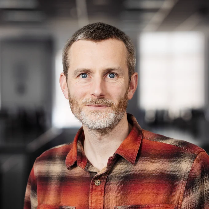 Dr Oliver Burton pictured wearing a red and brown plaid shirt standing in a softly lit indoor space with blurred background.