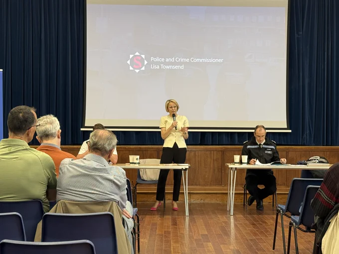 Image shows Surrey Police and Crime Commissioner in a yellow top holding a microphone standing in a hall behind a long trestle table.  She is speaking to an audience where we can see the backs of some audience members.  To her left is the Surrey Chief Constable Tim De Meyer who is in uniform reading some notes that are resting on the table.  Behind Lisa is a very large screen that has her name and role on it