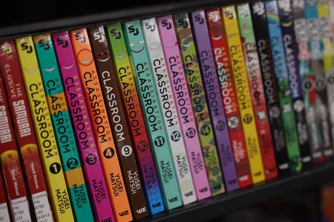 Close-up photos of manga and graphic novel shelves in a library collection, featuring titles including Hellsing, Demon Slayer, Attack on Titan, and Assassination Classroom.
