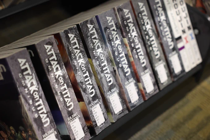 Close-up photos of manga and graphic novel shelves in a library collection, featuring titles including Hellsing, Demon Slayer, Attack on Titan, and Assassination Classroom.
