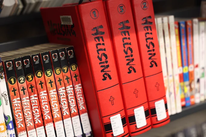 Close-up photos of manga and graphic novel shelves in a library collection, featuring titles including Hellsing, Demon Slayer, Attack on Titan, and Assassination Classroom.