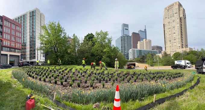 Workers planting greenery in a circular urban garden surrounded by tall city buildings under a cloudy sky.