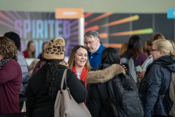 A group of people are gathered indoors, engaged in conversation. In the background, a colorful sign reads "Spirited" and there are different colored stripes emanating from the sign. The scene appears lively with attendees wearing winter clothing, suggesting a chilly season or location.